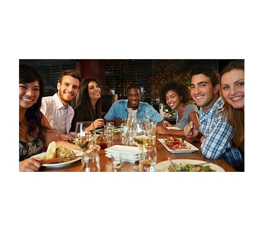 A group of seven people smiling and sitting around a restaurant table, celebrating Festival Week. They have plates of food and glasses of wine, enjoying the GBSB Festival 2024.