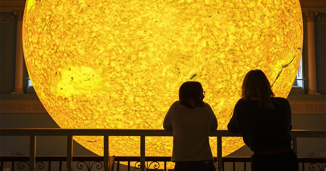 Two people stand at a railing during a Business Hive networking event, observing a large, illuminated model of the Sun indoors.